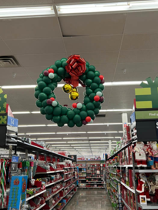 Christmas style balloon wreath that is green with small balloons in red, cranberry, and mint. A large red bow on the top finishes the design.